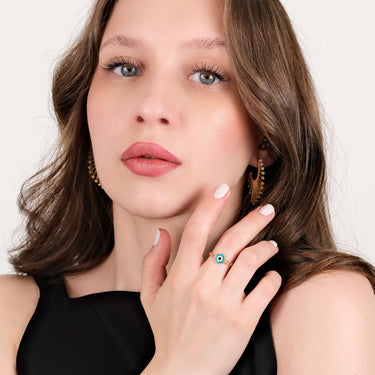 A woman with light brown hair and blue eyes wearing a black top, posing with her hand near her face to display a gold chain ring featuring a hexagonal evil eye design