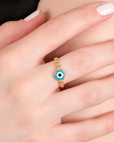 A close-up view of a hand with neatly manicured nails painted white, showcasing a gold chain ring with a hexagonal blue and white evil eye design.