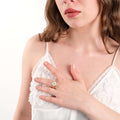 A close-up of a woman in a white lace top, her hand on her chest to show off a 14K solid gold ring with a daisy design.