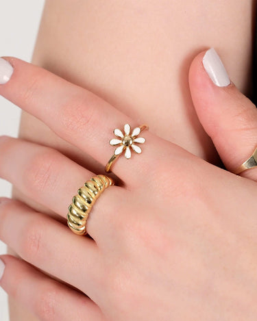 Close-up of a hand wearing a gold flower ring with white petals, alongside two other gold rings, against a neutral background.