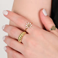 Close-up of a hand wearing a gold flower ring with white petals, alongside two other gold rings, against a neutral background.