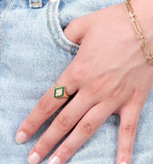 A close-up of a hand with a gold snake signet ring featuring a green and white enamel snake design, resting on the pocket of light blue denim jeans. The hand also wears a delicate gold chain bracelet.