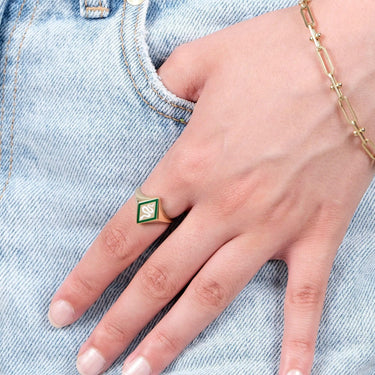 A close-up of a hand with a gold snake signet ring featuring a green and white enamel snake design, resting on the pocket of light blue denim jeans. The hand also wears a delicate gold chain bracelet.