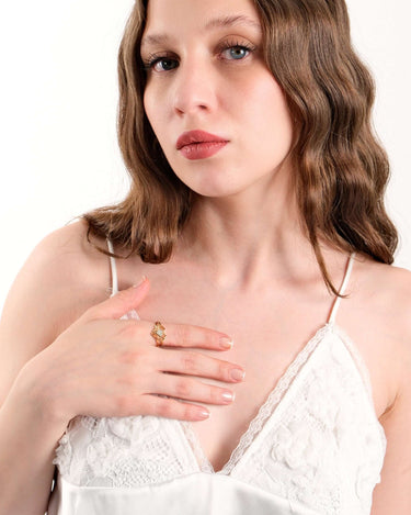 A woman in a white lace top resting her hand on her chest, highlighting a gold heart ring on her finger.