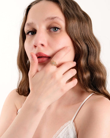 Close-up of a woman with wavy brown hair and blue eyes, touching her chin and displaying a gold heart ring on her finger.