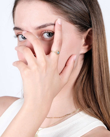 A young woman with straight light brown hair is posing with her hand near her face, showing off a gold evil eye ring with a blue gemstone.