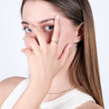 A young woman with straight light brown hair is posing with her hand near her face, showing off a gold evil eye ring with a blue gemstone.