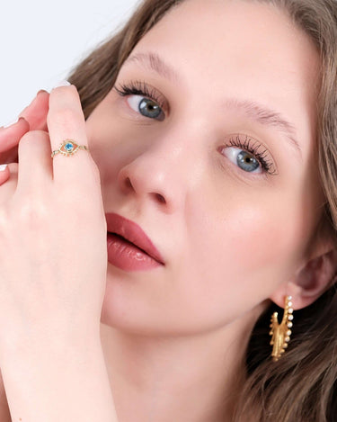 A close-up of a woman with light blue eyes and wavy light brown hair. She is wearing a gold evil eye ring with a blue gemstone on her index finger, held close to her face.