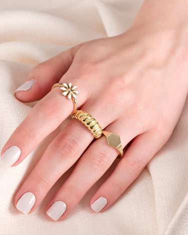 Close-up of a hand on a white fabric, wearing a gold flower ring with white enamel petals, and two additional gold rings.