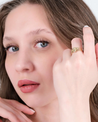 Woman with light makeup and wavy brown hair holding her hand near her face, displaying a gold bee ring with a honeycomb pattern.