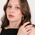 A close-up of a woman with light brown hair and blue eyes, wearing gold earrings and a gold chain ring with a hexagonal evil eye design, posing with her hand near her face.