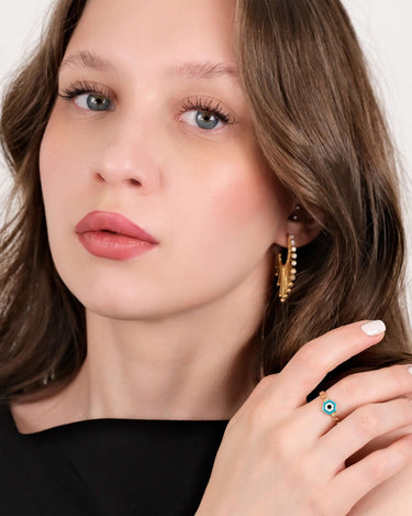 A close-up of a woman with light brown hair and blue eyes, wearing gold earrings and a gold chain ring with a hexagonal evil eye design, posing with her hand near her face.