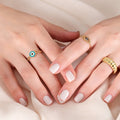 A pair of hands with white manicured nails, wearing multiple rings, including a gold chain ring with a hexagonal blue and white evil eye design.