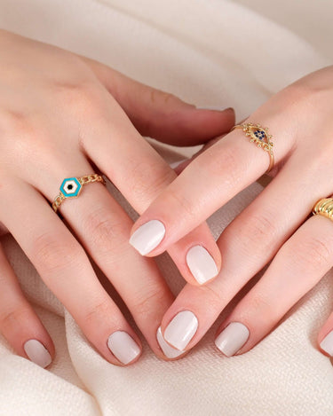 A pair of hands with white manicured nails, wearing multiple rings, including a gold chain ring with a hexagonal blue and white evil eye design.