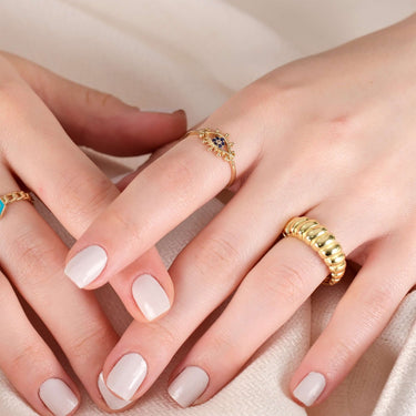 A close-up of two hands with manicured nails, one wearing a gold evil eye ring with blue stones and the other wearing a gold twisted ring.