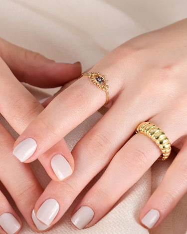 A close-up of two hands with manicured nails, one wearing a gold evil eye ring with blue stones and the other wearing a gold twisted ring.