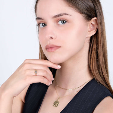 A woman with straight hair, wearing a gold evil eye ring and gold necklaces, posing with her hand near her chin.