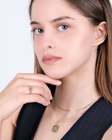 A woman with straight hair, wearing a gold evil eye ring and gold necklaces, posing with her hand near her chin.