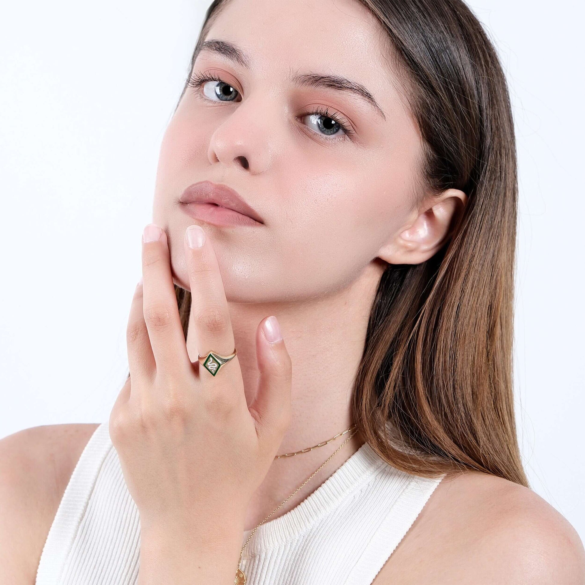 A close-up of a woman&#39;s face, looking directly at the camera, with her hand touching her chin. She is wearing a gold snake signet ring with a green and white enamel snake design and a delicate gold chain necklace.