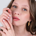 A close-up of a person with long brown hair and blue eyes, wearing a gold ring adorned with gemstones, a Mionza Chain Ring in 14K solid gold, and gold hoop earrings. They touch their face gently with manicured hands, one against their cheek and the other near their chin, showcasing the elegant jewelry.