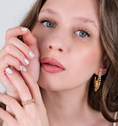 A close-up of a person with long brown hair and blue eyes, wearing a gold ring adorned with gemstones, a Mionza Chain Ring in 14K solid gold, and gold hoop earrings. They touch their face gently with manicured hands, one against their cheek and the other near their chin, showcasing the elegant jewelry.