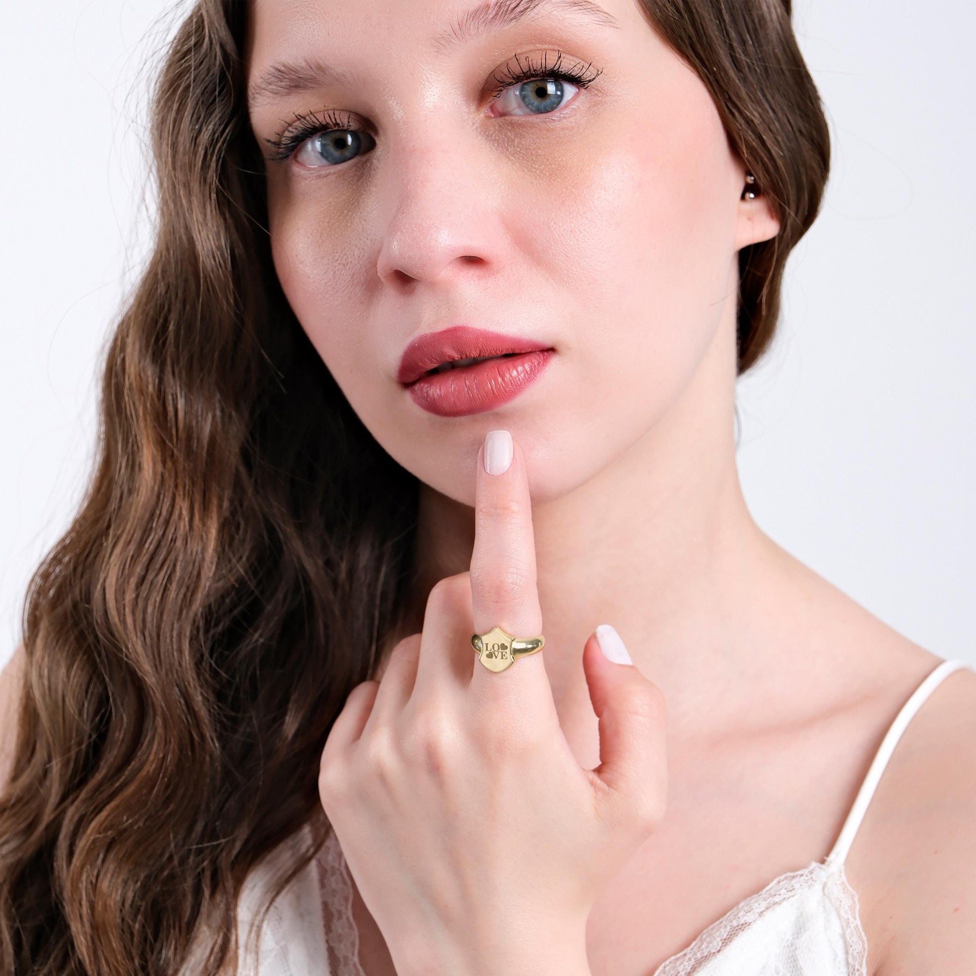 A young woman with wavy brown hair posing in a white lace top, touching her lips with her finger to highlight the 14K solid gold initial ring with &quot;LOVE&quot; engraving.