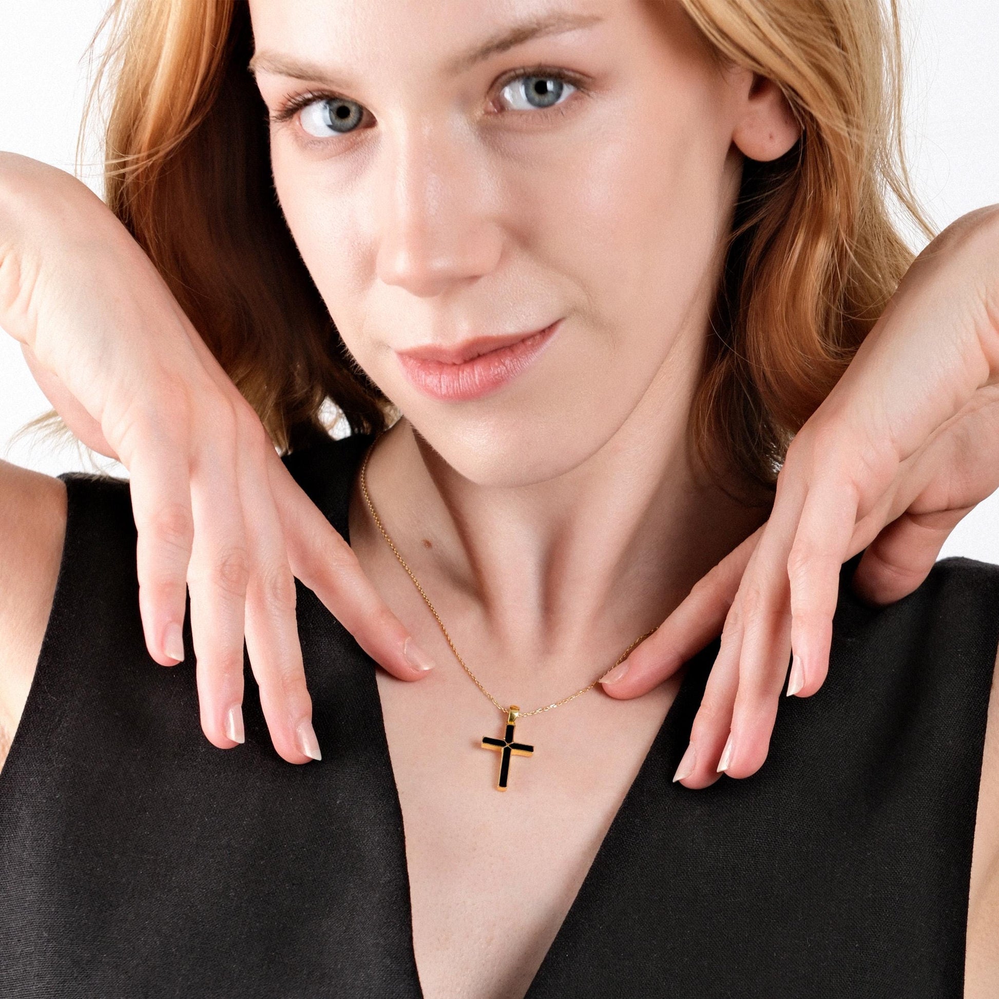 Woman gently holding a 14K gold vermeil necklace with a black cross pendant against her chest, highlighting the necklace&#39;s design.