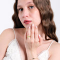 A young woman with long wavy brown hair wearing a white lace top, gently touching her face with her hand, displaying a 14K solid gold ring with the word &quot;LOVE&quot; engraved on a shield-shaped face.