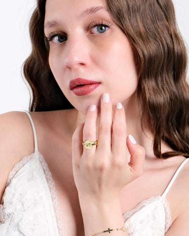 A young woman with long wavy brown hair wearing a white lace top, gently touching her face with her hand, displaying a 14K solid gold ring with the word &quot;LOVE&quot; engraved on a shield-shaped face.