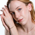 A woman with light skin and light brown hair gazes softly at the camera, her hands gently holding her wrist, which is adorned with a 14K solid gold onyx rosary bracelet.