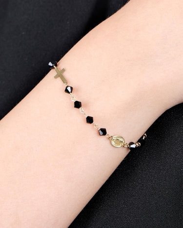 A close-up of a woman&#39;s wrist wearing a delicate 14K gold bracelet with black onyx beads, featuring a small cross charm and a medallion.