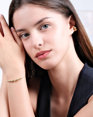 A woman with long brown hair and fair skin wearing an 18K Gold Vermeil beaded bracelet on her wrist, with her hands gently resting against her face.