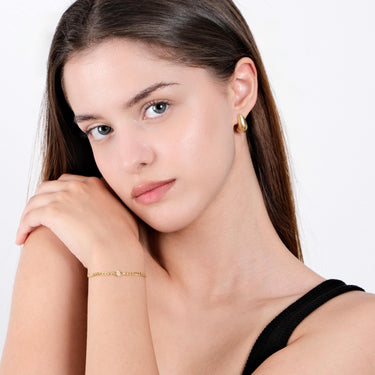 A woman with light skin and straight hair poses with her hand near her face, showcasing an 18K Gold Vermeil Evil Eye Curb Chain Bracelet and gold earrings, highlighting the bracelet&#39;s dainty design.