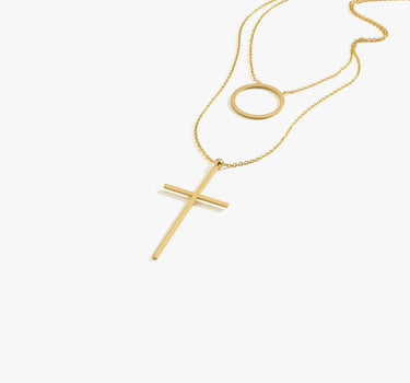 Product shot of an 18K gold vermeil layered necklace displayed flat, showcasing a minimalist cross pendant and a circular pendant on fine gold chains.