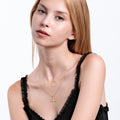 A young woman with light skin and straight blonde hair models an 18K gold vermeil layered cross necklace, wearing a black ruffled top against a plain background.