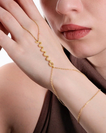 Close-up of a woman&#39;s hand resting on her cheek, showcasing a delicate gold snake hand chain bracelet draped across her hand, with a zigzag design as the central feature.