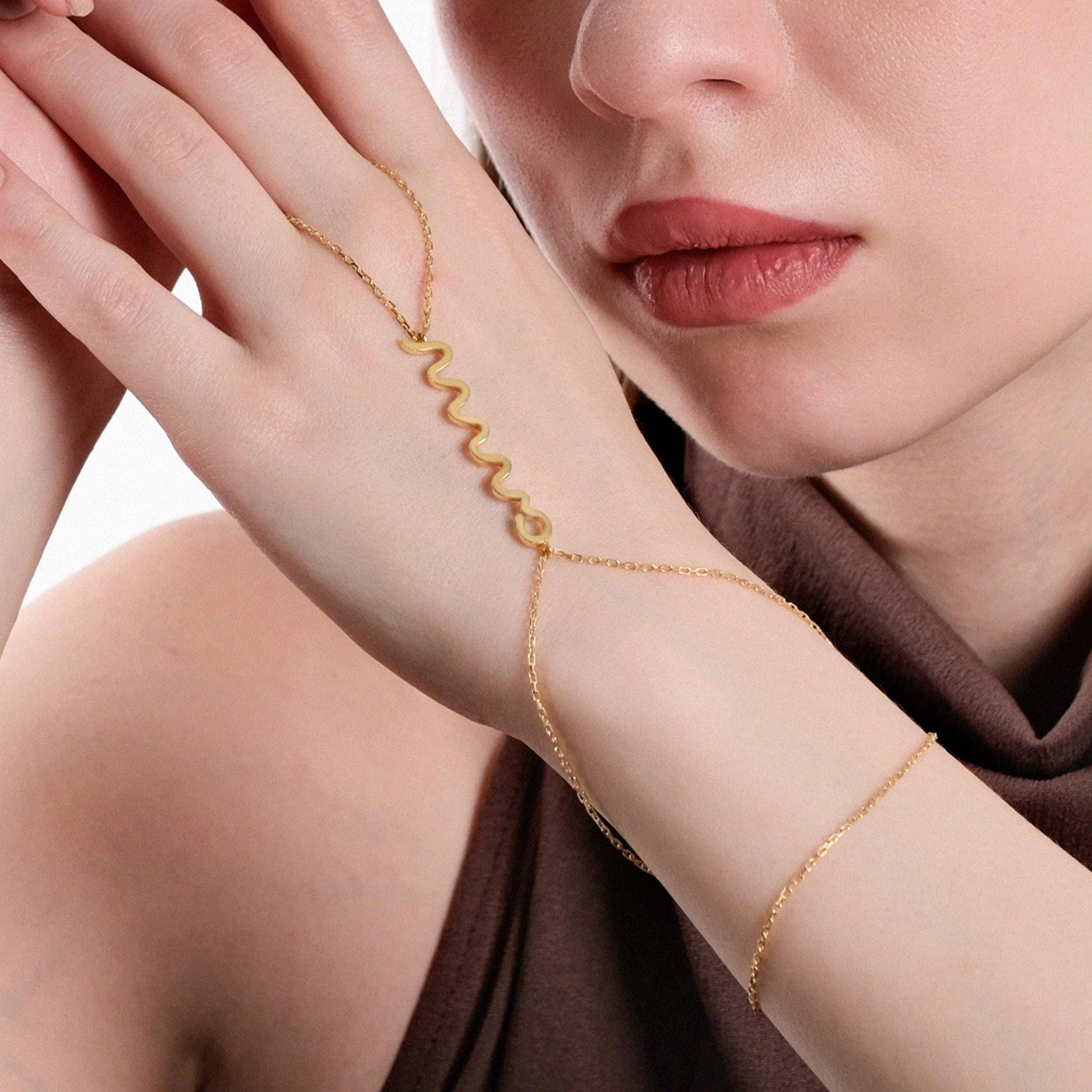 Close-up of a woman&#39;s hand resting on her cheek, showcasing a delicate gold snake hand chain bracelet draped across her hand, with a zigzag design as the central feature.