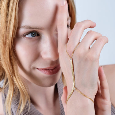 Woman with blonde hair gently touching her face with her right hand, which is adorned with a gold vermeil herringbone hand chain bracelet.