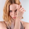 Woman with blonde hair gazing at the camera, wearing a gold vermeil herringbone hand chain bracelet on her right hand, which is held up near her face.