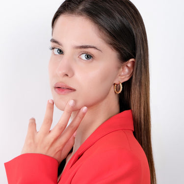 A woman in a red blazer, with long brown hair, wearing a medium-sized twisted hoop earring in 18K gold vermeil, looking at the camera with her hand near her face.