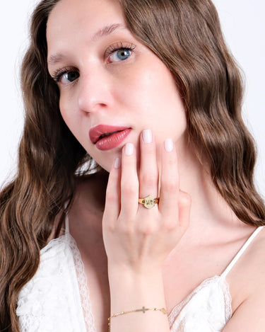 Close-up of a woman with wavy brown hair wearing a white lace top, holding her hand near her face to showcase a 14K solid gold initial ring with a &quot;LOVE&quot; engraving.