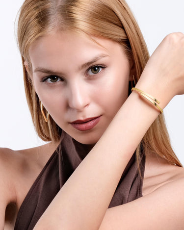 A woman with a focused expression shows off the 18K gold vermeil hinged bangle bracelet on her wrist, highlighting its elegant simplicity.