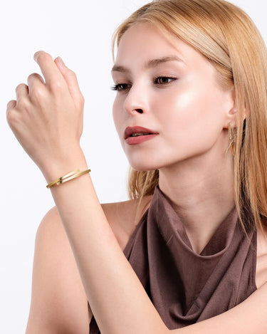 A woman with blonde hair models an 18K gold vermeil hinged bangle bracelet on her wrist, complementing her brown halter top and matching earrings.