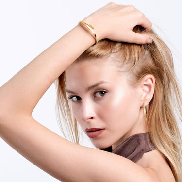 A woman poses with her hand on her head, revealing the 18K gold vermeil hinged bangle bracelet on her wrist, accentuating her sophisticated style.