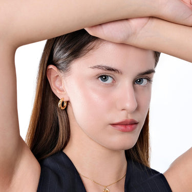 A front view of a woman with light skin and long, brown hair wearing an 18K gold vermeil chunky hoop earring, her arm raised, emphasizing the earring&#39;s bold, polished look with her simple black top.