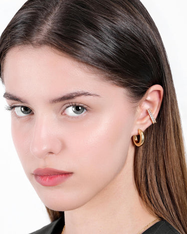 A close-up portrait of a woman with light skin and dark brown hair, wearing an 18K gold vermeil chunky hoop earring along with a thin, sparkling ear cuff.