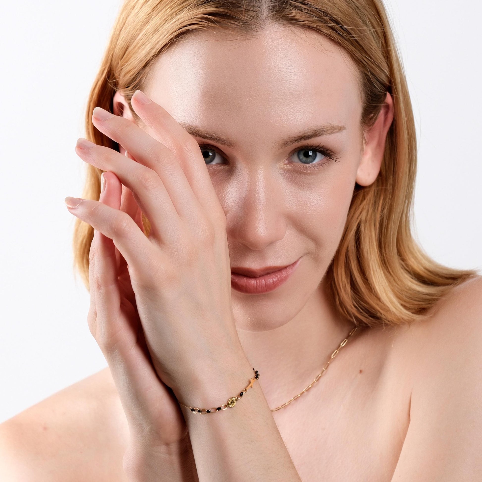 A woman with light skin and light brown hair smiles subtly, her hands framing her face and wrist adorned with a 14K solid gold onyx rosary bracelet. The bracelet, featuring a gold cross and medallion.