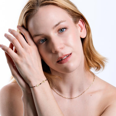 A woman with light skin and blonde hair softly smiles as she holds her wrist, showcasing a 14K solid gold onyx rosary bracelet with a cross and medallion charm.