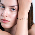 A close-up of a woman&#39;s face with light blue eyes, partially obscured by her arm, showcasing a 14K gold bracelet with black onyx beads, a cross charm, and a small medallion.