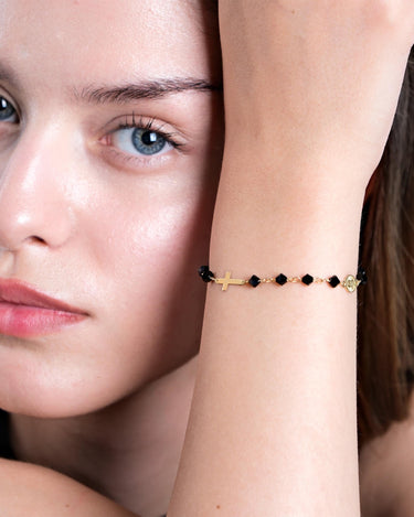 A close-up of a woman&#39;s face with light blue eyes, partially obscured by her arm, showcasing a 14K gold bracelet with black onyx beads, a cross charm, and a small medallion.